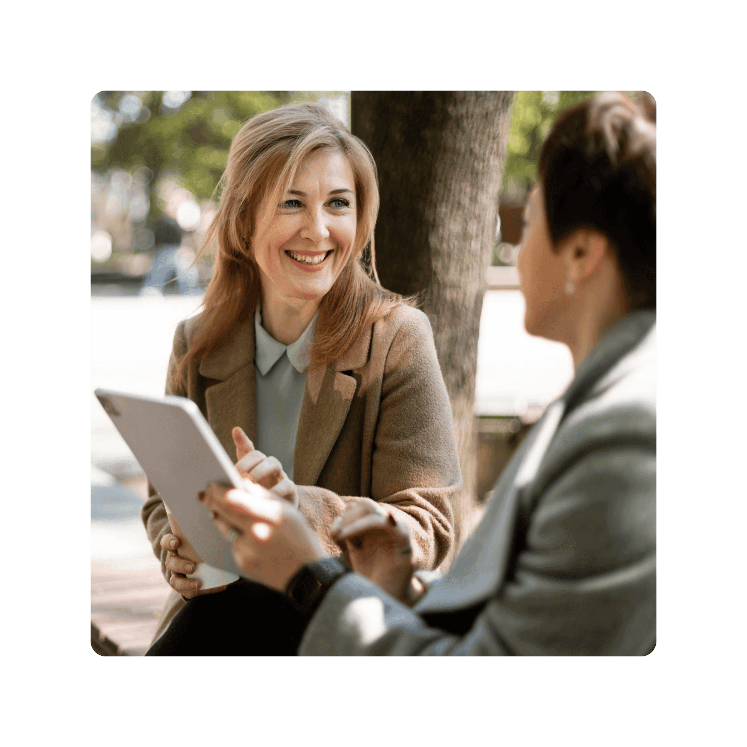 A woman shows another woman her tablet device on a bench